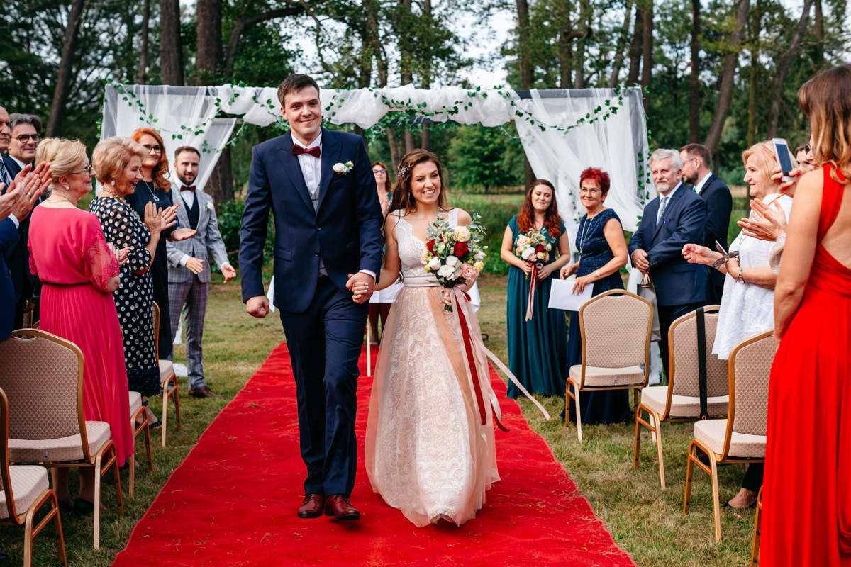 Ślub plenerowy w dworze Many. PAra Młoda idąca od ołtarza po czerwonym dywanie.